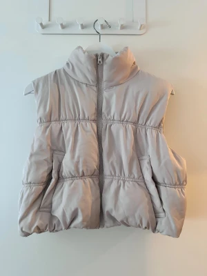 Beige pufferväst med dragkedja - Säljer en beige pufferväst med hög krage och dragkedja framtill. Västen har en quiltad design och två fickor på sidorna. Perfekt att slänga över en hoodie för en chill och snygg look.