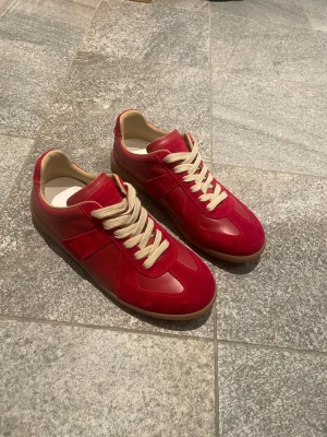 Maison Margiela röda sneakers - Maison Margiela sneakers i rött skinn och mocka med beige snörning och gummisula. Klassisk rund tå och platt sula, med snygga paneler och retrovibe. Perfekt för dig som gillar statement-färg och stilrena detaljer.
