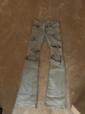 Blå lågmidjade jeans med hål - Säljer ett par blå jeans med flera slitningar och hål på framsidan för en edgy look. Baktill har de två stora mörkblå bakfickor som kontrasterar mot resten av byxan. Modellen är bootcut och materialet är klassisk denim. 