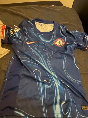 Chelsea FC blå Nike fotbollströja - Chelsea FC matchtröja från Nike i blått med unikt abstrakt mönster i ljusblå och vita toner. Kortärmad modell med rund hals och Dri-FIT-material som andas. Klubbmärke och Nike-logga på bröstet, samt Premier League-detaljer på ärmen.