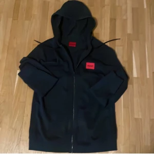 Svart hoodie från Hugo med röd logga - Svart hoodie från Hugo med röd patch och svart text på bröstet. Tröjan har huva med snörning, hel dragkedja framtill och långa ärmar. Materialet är mjuk bomull och passformen är normal. Perfekt för chill dagar och streetwear.