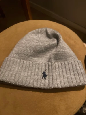 Grå stickad mössa från Polo Ralph Lauren har aldrig använt  - Snygg grå stickad mössa från Polo Ralph Lauren med klassisk ribbad kant och broderad marinblå logga framtill. Perfekt för kyliga dagar och enkel att matcha med olika outfits.