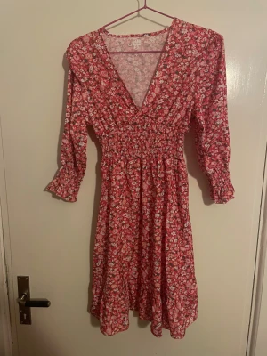 Rosa blommig klänning med volang - Supersöt rosa klänning med blommigt mönster, v-ringad och smockad midja. Långärmad med volangdetaljer vid ärmslut och nederkant. Perfekt för dig som gillar romantisk stil och vill ha något som sticker ut lite extra.