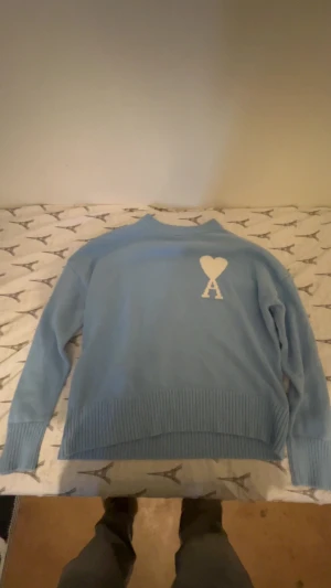 Ljusblå stickad tröja från Ami - Snygg ljusblå stickad tröja från Ami Alexandre Mattiussi med vit hjärt- och A-logga på bröstet. Tröjan har ribbade muddar vid ärmslut och nederkant samt rund hals. Perfekt för dig som gillar stilrena detaljer och vill ha något som sticker ut.