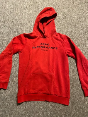 Röd hoodie från Peak Performance - Riktigt snygg röd hoodie från Peak Performance med svart broderad logga på bröstet. Klassisk känguruficka framtill, huva och ribbade muddar. Perfekt för chill dagar eller när du vill ha en sportig look. Storlek 170.