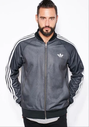 Adidas Originals träningsjacka 2xl - Snygg Adidas Originals träningsjacka i mörkgrå/svart med diskret diamantmönster och klassiska vita tre ränder längs ärmarna. Ikoniskt Adidas-Trefoil-tryck på bröstet och dragkedja i guld-look som ger en riktigt cool retrostil.  Storlek: 2XL Perfekt skick  En modell som inte längre finns i butik och kan vara svår att få tag på – perfekt både för samlare och för dig som vill sticka ut med en unik Adidas-look!