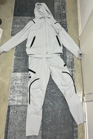 Ljusgrå träningsset från Under Armour - Säljer ett ljusgrått träningsset från Under Armour med svarta detaljer. Setet består av en hoodie med dragkedja och matchande byxor, båda med flera svarta dragkedjor och sportiga linjer. Perfekt för dig som gillar stilren och funktionell träningsoutfit.