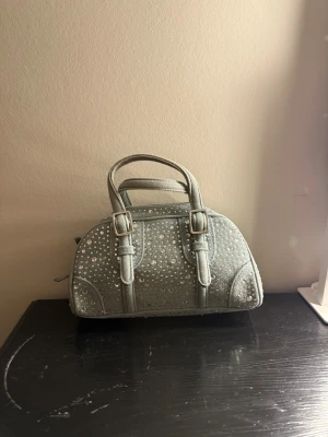 Silver handväska med paljetter - Snygg handväska i silverfärgat tyg med glittrande paljetter över hela väskan. Väskan har två handtag
