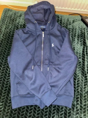 Marinblå hoodie från Polo Ralph Lauren - Snygg marinblå hoodie från Polo Ralph Lauren med dragkedja och klassisk logga på bröstet. Tröjan har huva med snörning, ribbade muddar och två fickor framtill. Tillverkad i mjuk bomullsmix som är skön mot huden. Perfekt för chill dagar och streetwear.