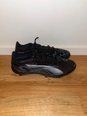 Svarta Puma fotbollsskor - Säljer ett par svarta fotbollsskor från Puma med snygga blå linjer och silvriga Puma-loggor på sidorna. Skorna har snörning och platt sula med dobbar för gräsplan. Ovandelen är i syntetmaterial och designen är riktigt sportig och modern.