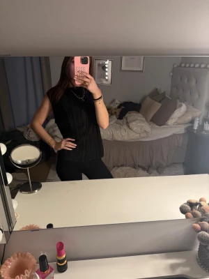 Svart ribbad ärmlös topp - Säljer en svart ärmlös topp med ribbad struktur och rak passform. Toppen har en enkel, stilren look med rund hals och är tillverkad i ett mjukt syntetmaterial som känns skönt mot huden. Perfekt att styla med smycken och jeans för en clean vibe.