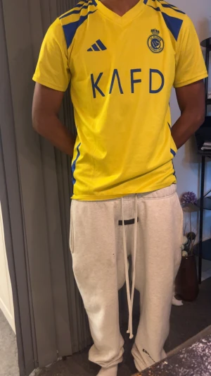Al Nassr Ronaldo 7 gul fotbollströja - Gul Al Nassr fotbollströja från Adidas med blå detaljer och tryck. Framsidan har klubbmärket och KAFD-logga, baksidan har Ronaldo och nummer 7 i blått. Tröjan är kortärmad och gjord i lätt, ventilerande material som passar perfekt för träning eller match.