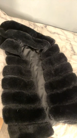 Svart pälsjacka med bred krage - Säljer en svart pälsjacka med bred krage och fluffiga paneler. Jackan har en lyxig känsla och är perfekt för dig som vill sticka ut med en cool och trendig look. Materialet är mjukt och ger en varm vibe under kalla dagar.