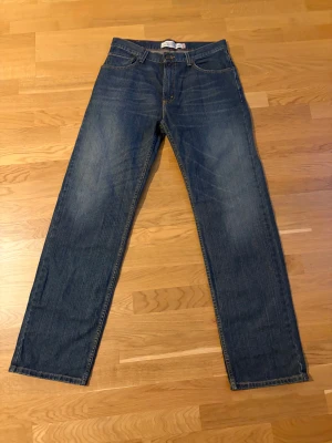 Levi's 501 blå jeans straight fit - Klassiska Levi's 501 jeans i blå tvätt med raka ben och normal passform. Jeansen har fem fickor och ikonisk läderpatch bak i midjan. Tillverkade i slitstarkt jeanstyg med snyggt slitage vid benslut. Perfekta för en chill och tidlös look.