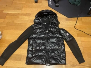 Svart Monclercardigan med huva - Säljer en svart cardigan från Moncler med glansig finish och stickade svarta ärmar. Jackan har huva, dragkedja framtill och Moncler-logga på ärmen. Insidan har en cool serietidningsetikett. Perfekt för dig som vill ha en snygg och unik jacka med street vibe. Skriv om ni har frågor, pris kan diskuteras.