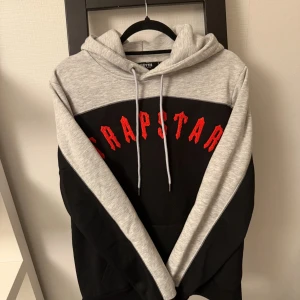 Trapstar hoodie i grått och svart - Snygg hoodie från Trapstar i grått och svart med röd broderad logga över bröstet. Tröjan har huva med dragsko och en klassisk loose passform. Perfekt för dig som gillar streetwear och vill sticka ut med en fet logga.