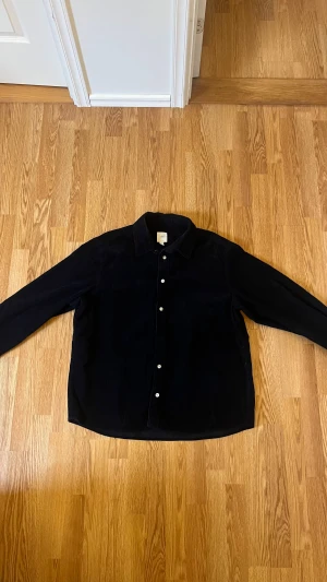 Svart manchesterskjorta från H&M L.O.G.G - Svart skjorta i mjuk manchester från H&M L.O.G.G, storlek XL. Klassisk krage och vita knappar framtill. Skjortan har lång ärm och en loose passform som ger en chill vibe. Perfekt för dig som gillar stilrena och bekväma plagg.