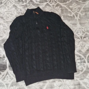 Polo Ralph Lauren Quarter - Zip - Svart kabelstickad tröja från Polo Ralph Lauren med halv dragkedja och brunt läderdetalj på zip. Röd broderad logga på bröstet. Hög krage och ribbade muddar. Perfekt för dig som gillar klassisk stil med en modern touch.