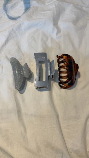 Tre hårklämmor i olika färger - Säljer ett set med tre hårklämmor: en glittrig silverklämma med mjuk form, en stilren rektangulär klämma i blank silverfärg och en klassisk brun klämma med sköldpaddsmönster. Perfekt för att fixa frisyren snabbt och snyggt.