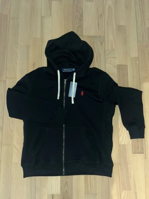 Svart hoodie från Polo Ralph Lauren - Svart hoodie från Polo Ralph Lauren med vit snörning i huvan och röd logobrodyr på bröstet. Tröjan har dragkedja framtill, två fickor och ribbade muddar vid ärmslut och nederkant. Perfekt för chill dagar och streetstyle.