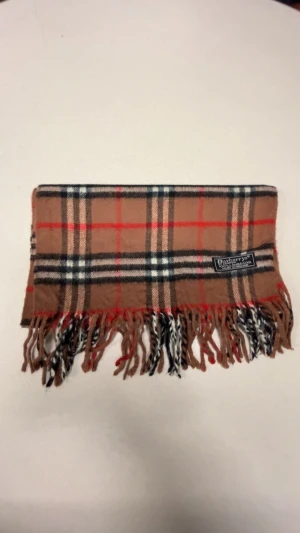 Rutig halsduk från Burberry i ull - Snygg halsduk från Burberry med klassiskt rutigt mönster i beige, svart, vitt och rött. Gjord i mjuk ull och har fransar i ändarna. Perfekt accessoar för att lyfta din outfit med lite lyxig London-vibe.