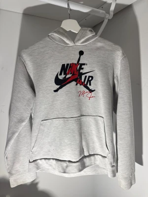 Grå Nike Air hoodie med tryck - Snygg grå hoodie från Nike Air med stort svart och rött tryck på bröstet och Michael Jordan-signatur. Tröjan har huva, lång ärm och en klassisk magficka. Perfekt för dig som gillar streetwear och sportig stil.
