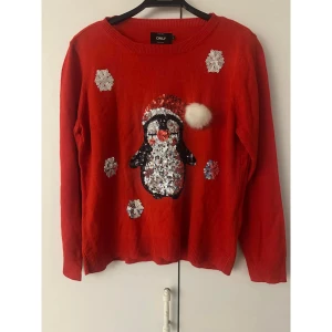 Röd jultröja med paljetter från ONLY - Röd stickad tröja från ONLY med ett gulligt paljettmönster av en pingvin i tomteluva och snöflingor. Tröjan har lång ärm och rund hals. Perfekt för att maxa julstämningen med lite extra glitter och fluffig detalj på luvan. Tröjan är en tunn i tyget. Liten i storleken.