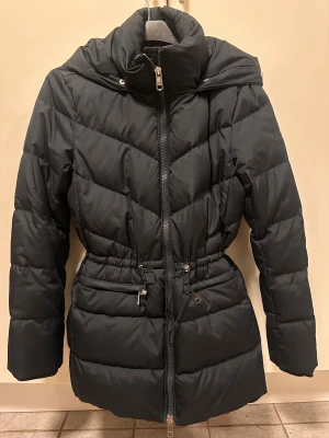 Marinblå  dunjacka från Tommy Hilfiger - Marinblå dunjacka från Tommy Hilfiger med huva, quiltad design och snygga snörningsdetaljer i midjan. Jackan har dragkedja framtill, långa ärmar och ett broderat H-märke på armen. Perfekt för kalla dagar och har en modern, lite längre passform.