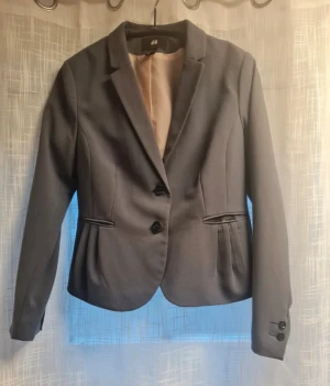 Grå kavaj från H&M, storlek 44 - Snygg grå kavaj från H&M i storlek 44. Klassisk modell med två knappar framtill, figursydd passform och diskreta fickor. Fodret är ljusrosa med små prickar. 