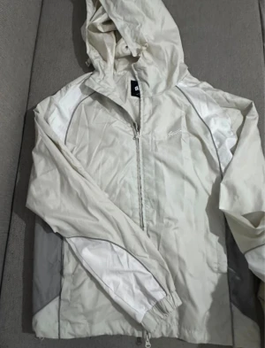 Vit vindjacka från SWD, storlek L - Säljer en stilren vit vindjacka från Stayd i storlek L. Jackan har huva, dragkedja framtill och coola detaljer i vitt och grått. Perfekt för blåsiga dagar och enkel att matcha med allt. Tillverkad i lätt polyester för skön känsla.