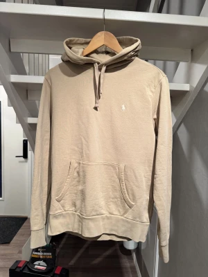 Beige hoodie från Polo Ralph Lauren - Använd 1 gång, som en helt ny hoddie. Snygg beige hoodie från Polo Ralph Lauren med klassisk broderad logga på bröstet. Tröjan har huva med snörning, känguruficka och långa ärmar. Tillverkad i mjuk bomull för skön komfort och stilren look.
