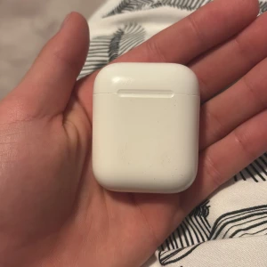 AirPods 2 - Apple AirPods 2 med laddningsetui. Trådlösa hörlurar med enkel anslutning till iPhone och andra Apple-enheter. Skick: synligt slitage och repor på etuiet, samt smuts på insidan av locket. Hörlurarna fungerar och laddas som de ska.