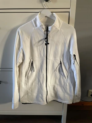 Vit Tröja från C.P. Company - Snygg vit tröja från C.P. Company med svart dragkedja och coola snörningsdetaljer vid fickorna och ärmen. tröjan har hög krage, lång ärm och en rund svart logotyp-patch på vänster ärm. 