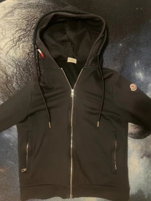 Svart hoodiejacka från Moncler - Svart hoodiejacka från Moncler med dragkedja framtill och två sidofickor med dragkedja. Jackan har en stor huva med snörning och Moncler-logga på vänster ärm. Snygga metalländar på snörena och stilren design, perfekt för chill och streetwear. Passar även (S), Äkta och har NFC.