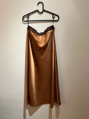 Brun satinlångkjol från Costa Mani - Säljer en snygg långkjol från Costa Mani i en glansig brun satin liknande material. Kjolen har ett mörkbrunt midjeband som ger en cool kontrast. Den är rak i modellen och har en mjuk, lyxig känsla. Perfekt för dig som gillar stilrena och trendiga plagg.