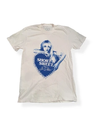 Sabrina Carpenter- T-shirt Short n' Sweet Tour 2025 - Vit t-shirt med blått tryck fram och bak från Sabrina Carpenters Short n' Sweet Tour 2025. Motiv med porträtt och hjärta, samt turnédatum på ryggen. Tillverkad i 100% bomull och har klassisk passform med korta ärmar. Endast tvättad.