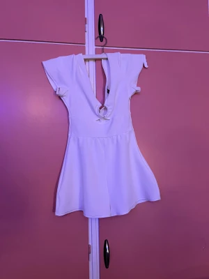 Vit kort klänning från Divided XS - Superfin vit kort klänning från Divided i storlek XS. Klänningen har djup v-ringning både fram och bak, korta ärmar och en snygg knyt-detalj framtill. Materialet är strukturerat och känns lätt och bekvämt. Perfekt för varma dagar när du vill sticka ut lite extra.