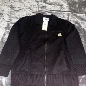 Svart stickad tröja från Carhartt - Svart stickad tröja från Carhartt med dragkedja framtill, klassisk krage och ribbade muddar. Tröjan har två snedställda fickor och en liten bröstficka med Carhartt-logga. Tillverkad i ekologisk bomull för extra skön känsla.