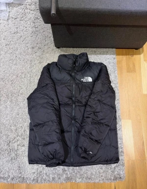 North face jacka - Säljer en svart pufferjacka från The North Face med broderad logga på bröstet. Jackan har hög krage, dragkedja framtill och elastiska muddar vid ärmslut. Perfekt för kalla dagar och har en clean, stilren look som funkar till allt.