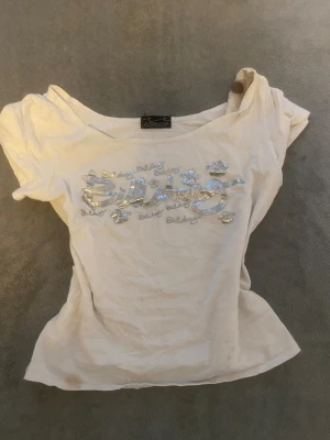 off shoulder/normal vit Billabong t-shirt med silvertryck - (den bruna fläcken är henna som torkade ut) Vit t-shirt från Billabong med korta ärmar och bred, något offshoulder halsringning. Framsidan har ett glansigt silvertryck med Billabong-logga och blommor. Materialet är mjuk bomull och passformen är slim. Perfekt för dig som gillar surfstil och streetwear.