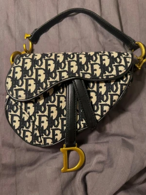  handväska med monogram - Snygg väska med ikoniskt svart och beige monogrammönster. Väskan har en unik sadelform, guldfärgade metalldetaljer. Detta är inte den stora modellen utan den mindre, den är perfekt för utgångar/middagar.