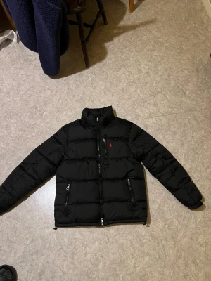 Svart pufferjacka från Polo Ralph Lauren - Svart pufferjacka från Polo Ralph Lauren med röd logga på bröstet. Jackan har hög krage, tre framfickor med dragkedja och quiltad design. Materialet är syntet och dun, perfekt för kalla dagar. Snygg och stilren look med långärmade ärmar och normal passform. Den är varm och bra att använda på vintern, den har inga skador.