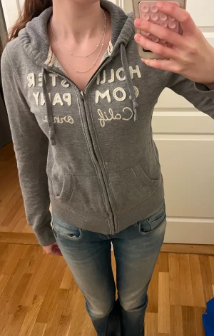 Grå hoodie från Hollister med dragkedja - Säljer en grå hoodie från Hollister med vit broderad text på bröstet. Tröjan har huva med snörning, två fickor fram och dragkedja hela vägen. Mjuk insida och klassisk passform. Storlek S. Säljer då den inte kommer till användning!💗 