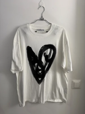 Vit Acne Studios t-shirt med svart hjärta i velour storlek M - Vit t-shirt från Acne Studios med ett stort svart hjärtmotiv på framsidan. Modellen är loose fit med korta ärmar och rund hals. Materialet är mjuk bomull och trycket har en cool, grafisk känsla som sticker ut.