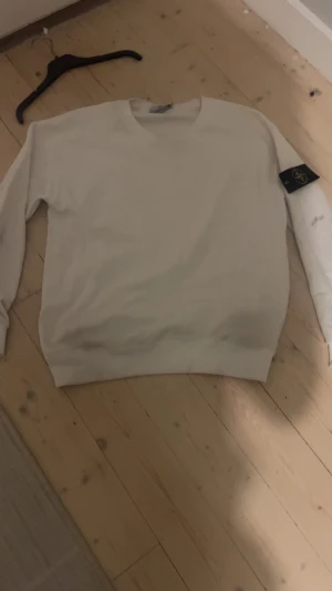 Stone Island sweatshirt - Vit sweatshirt från Stone Island med klassisk rund hals och ribbade muddar. Tröjan har lång ärm och den ikoniska svarta patchen med gult kompassmärke på vänster ärm. Materialet är mjuk bomull, perfekt för en clean och stilren look.