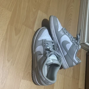 Nike Dunk Low vit/grå sneakers - Säljer ett par Nike Dunk Low sneakers i fräscht vitt och ljusgrått. Klassisk siluett med rund tå, snörning och platt sula. Ovandel i skinn och syntet, med tydlig Nike-logga på sidan och broderad logga bak. Perfekta för dig som gillar stilrena och cleana sneakers.