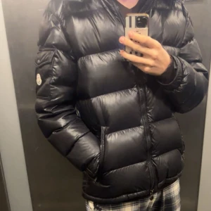 MONCLER MAYA  - Säljer en svart pufferjacka från Moncler med klassisk logga på ärmen. Jackan har en glansig finish, Kan gå ner i pris vid snabb äffär! Defekter finns inte mycket av enbart nån liten reva/hål om man ens kan kalla det för hål! Inget dun eller nått kommer ut, kom privat för frågor! Passar de med M på jackor och kanske L 