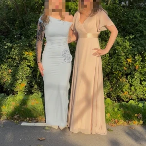 Balklänning  - Elegant långklänning i ljusbeige med v-ringad omlott-topp, korta fladdriga ärmar och brett satinband i midjan. Kjolen faller mjukt och materialet är lätt och flowy, perfekt för speciella tillfällen. Stilren och feminin look.