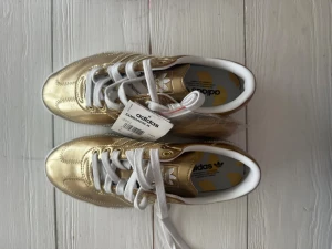 Adidas Sambarose guld sneakers - Adidas Sambarose W sneakers i metallic guld med vita detaljer och snörning. Skorna har en chunky plattformssula i vitt och klassiska Adidas-ränder på sidan. Helt nya med lapp kvar.