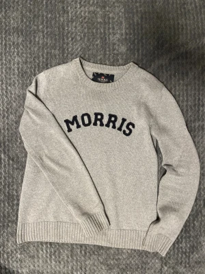 Morris grå stickad  - Snygg grå stickad tröja från Morris med svart broderad logga på bröstet och en liten brittisk flagga på ärmen. Rund hals och ribbade muddar vid ärmslut och nederkant. Perfekt för dig som gillar klassisk stil med en modern touch.  